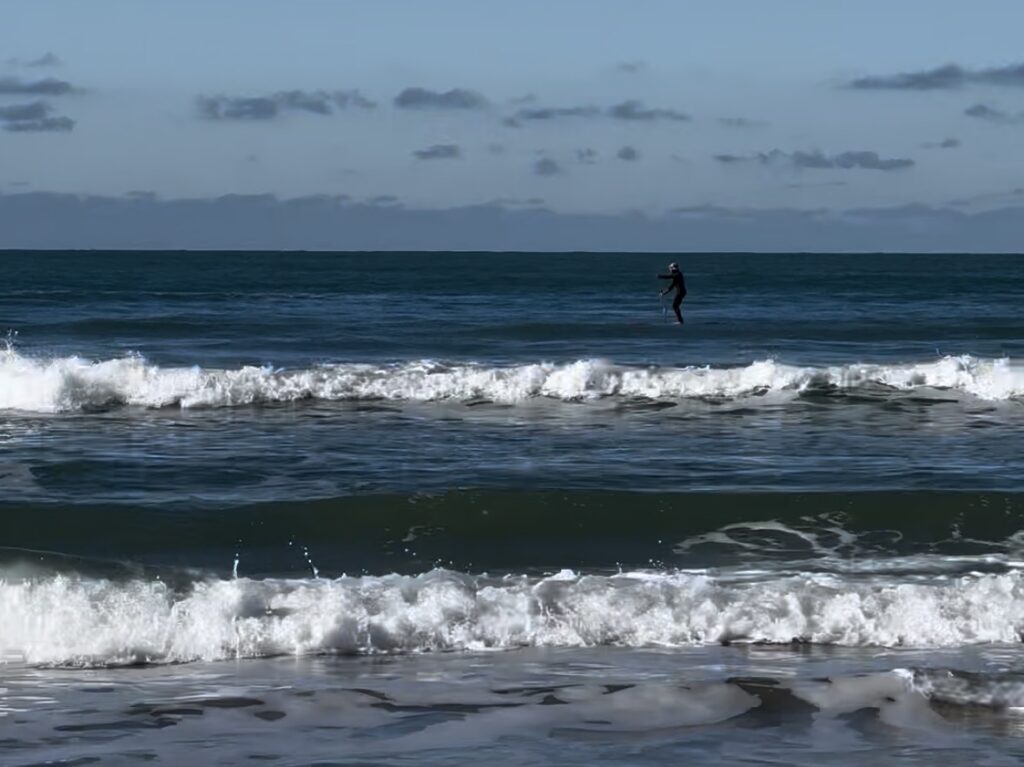 orilla de playa con oleaje y persona haciendo padel surf