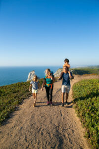 Active family couple and children hiking along seaside, walking on path. Boy riding on dads neck. Full length. Nature and recreation concept.